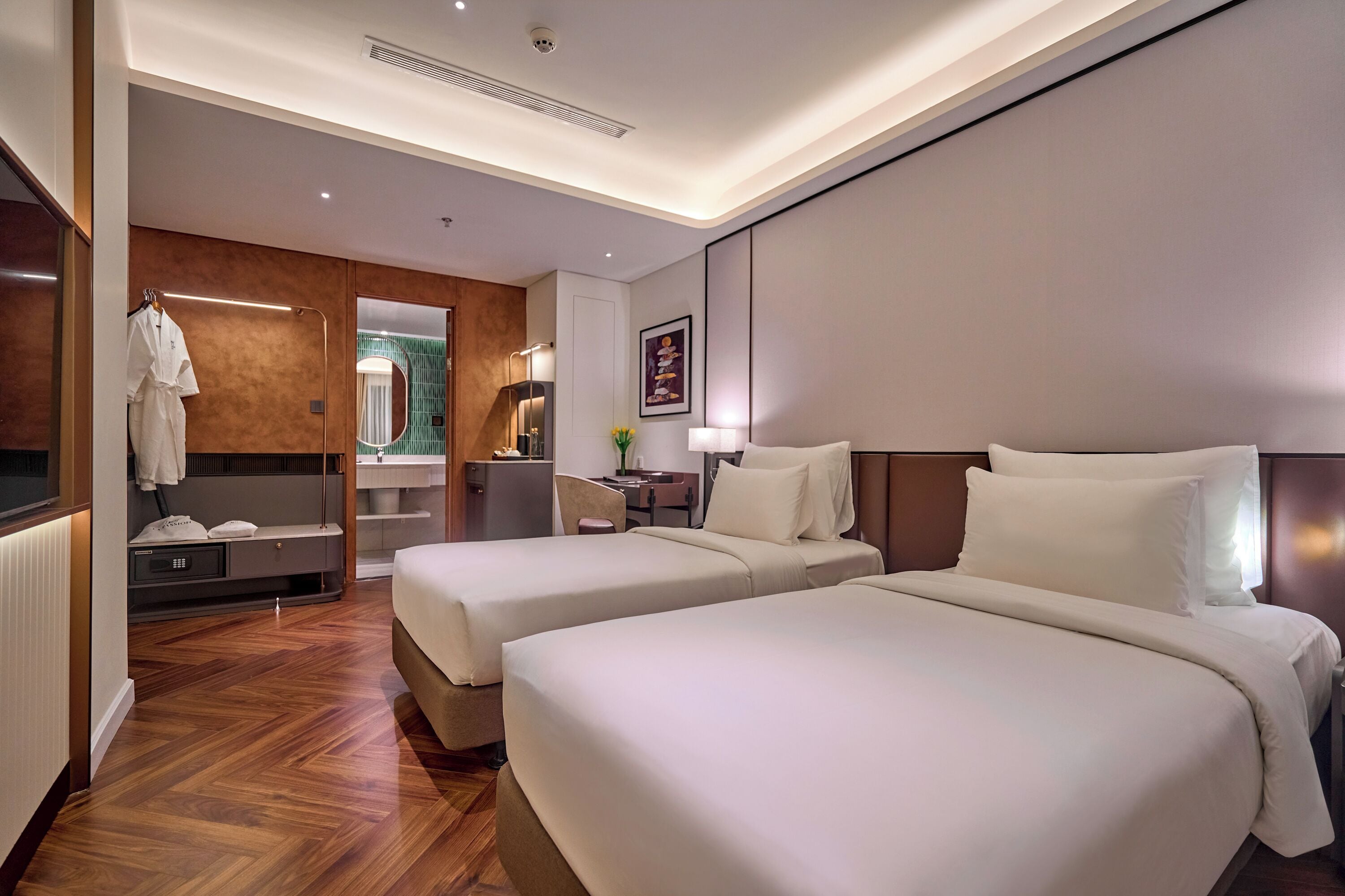 Une chambre d’hôtel avec deux lits, un plancher en bois, un miroir, un bureau et une salle de bain visible à travers une porte ouverte.