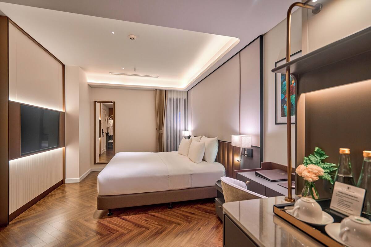 A modern hotel room with a large bed, a desk with a laptop, a mirror, and a vase with flowers.