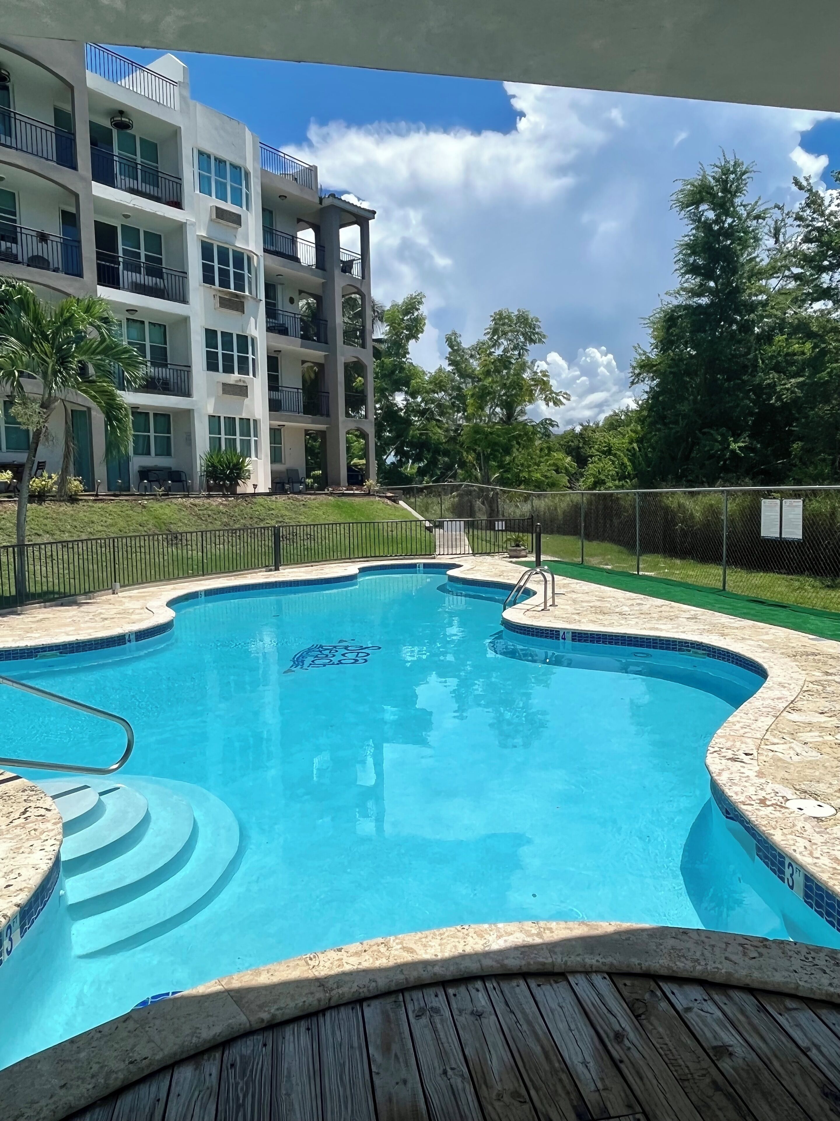 Piscina com deck de madeira, cercada por uma área delimitada, e um edifício com varandas ao fundo.