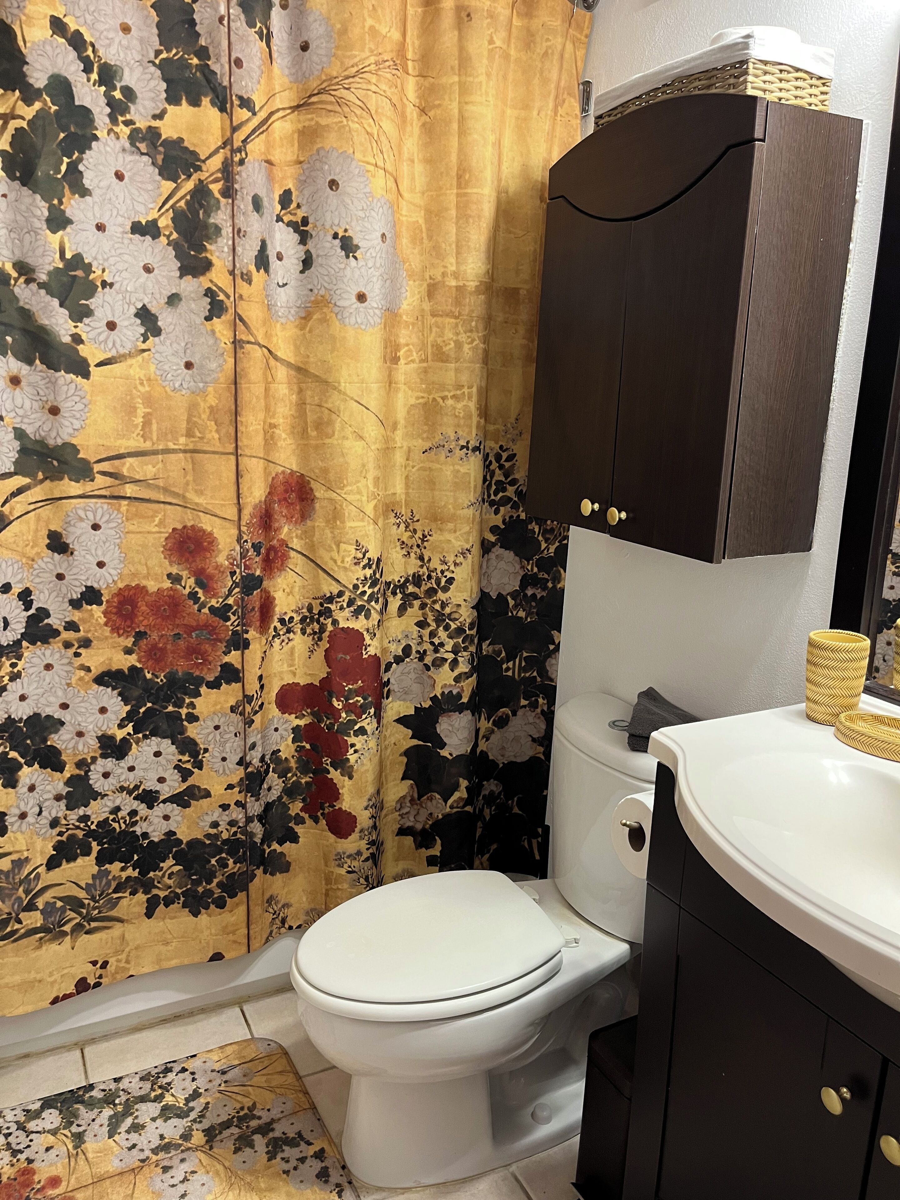 Un baño con cortina de ducha estampada con flores, un inodoro blanco y un mueble de madera oscura con lavamanos blanco.