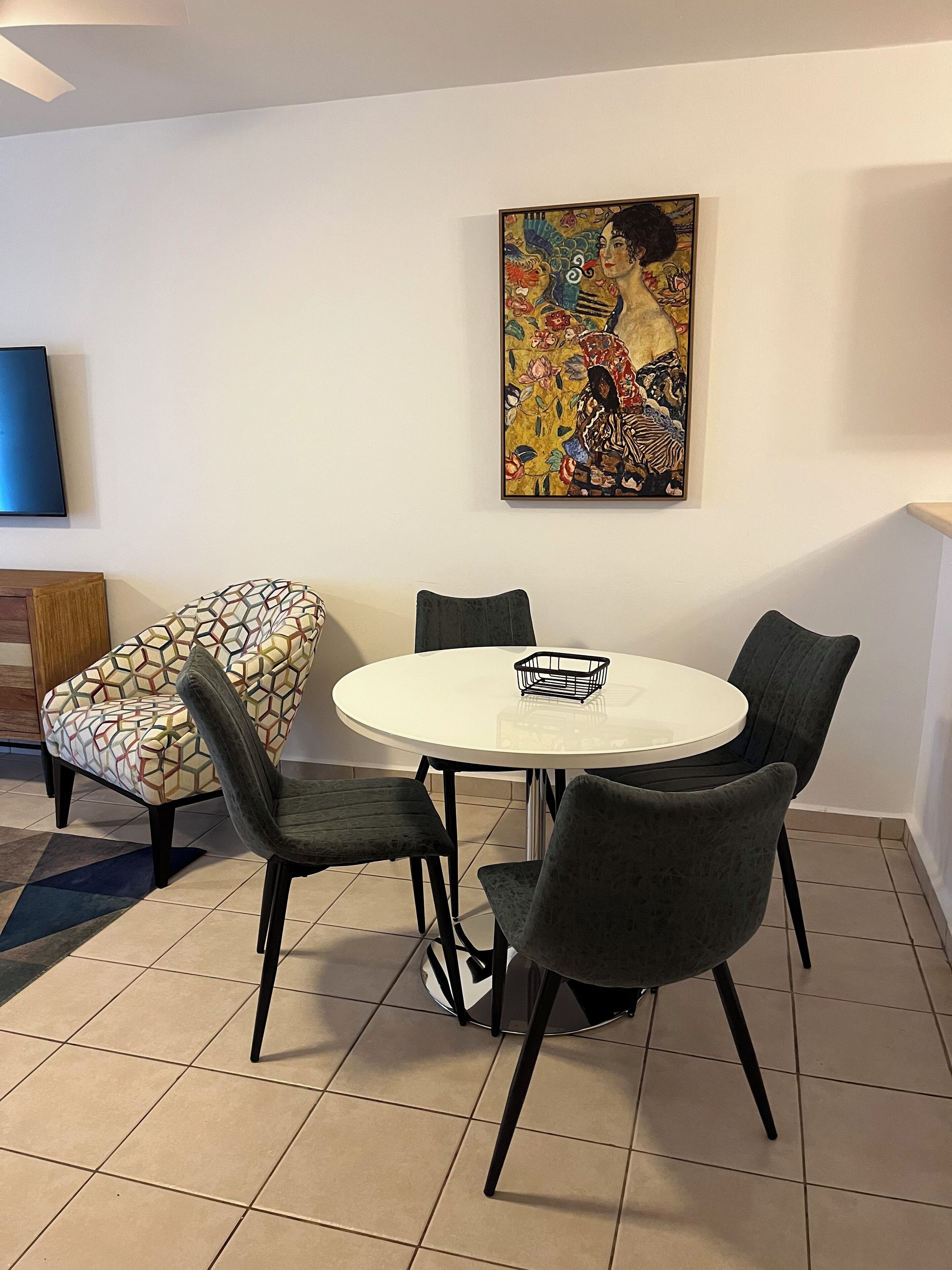 Een eetgedeelte met een ronde tafel, zes stoelen en een kleurrijk schilderij aan de muur.