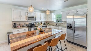 Una cocina moderna con armarios blancos, electrodomésticos de acero inoxidable, una isla de madera con taburetes altos e iluminación colgante.