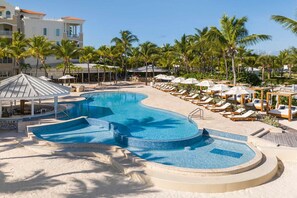 Uma área de piscina em resort, com espreguiçadeiras e guarda-sóis, cercada por palmeiras e um edifício com arquitetura mediterrânea.