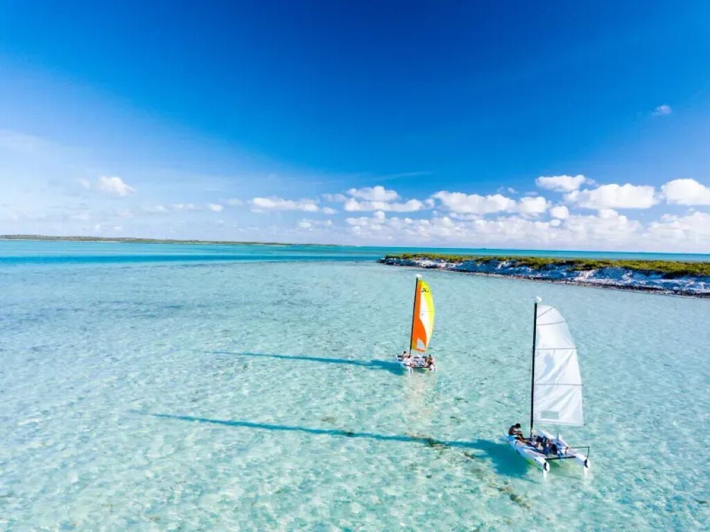 Une mer turquoise et limpide où voguent deux voiliers : l’un arbore une voile jaune et blanche, l’autre une voile blanche, le tout sous un ciel d’un bleu éclatant.