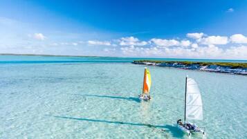 Une mer turquoise et limpide où voguent deux voiliers : l’un arbore une voile jaune et blanche, l’autre une voile blanche, le tout sous un ciel d’un bleu éclatant.