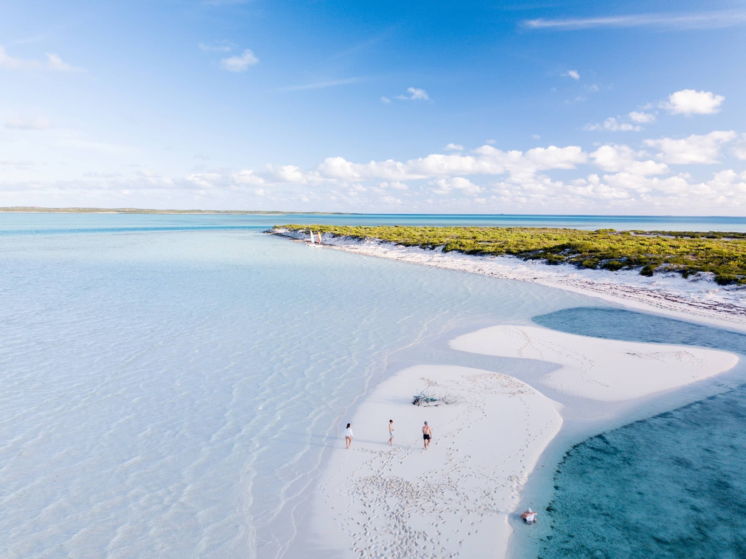 A beach with clear turquoise water, white sand, and sparse vegetation.