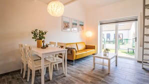 Un salon moderne avec une table à manger, des chaises blanches, un canapé jaune et une table basse. Des tableaux encadrés ornent le mur, et une grande fenêtre offre une vue sur l’extérieur.
