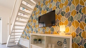 Un salon moderne doté d’un plafond en pente, d’un téléviseur à écran plat fixé au mur, d’une console blanche agrémentée d’objets décoratifs et d’un escalier dont le mur est orné d’un motif de feuilles.