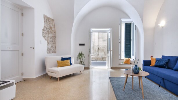 Una sala de estar amplia con un sofá blanco, un sofá azul, una mesa redonda y un amplio arco que da a un área exterior.