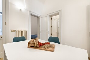 Ein Essbereich mit einem weißen Tisch, zwei blauen Stühlen, einem Holzschneidebrett mit Tomaten und Pasta sowie einer Papiertüte.