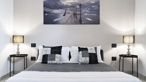 Un dormitorio con una cama, dos mesitas de noche, dos lámparas y un cuadro colgado en la pared que muestra un muelle.