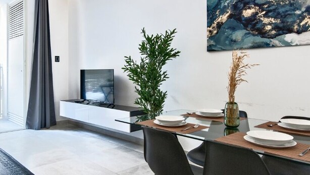 Una sala de estar moderna con un sofá, una mesa de comedor para cuatro personas, un televisor en un estante empotrado y un cuadro abstracto grande en la pared.