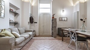 A living room with a sofa, a dining table with chairs, a kitchen area, and a hallway leading to a door. - Arco Barocco - Lecce Selection (Lecce)