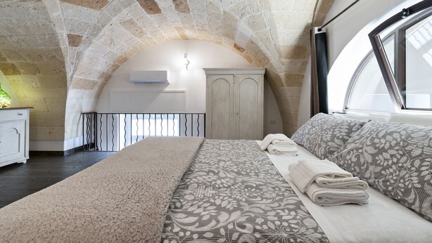 A bedroom with a stone ceiling, a bed with patterned bedding, a wardrobe, and a window with a view. - Arco Barocco - Lecce Selection (Lecce)