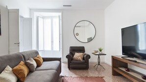 En moderne stue med en grå sofa, en brun lænestol, et rundt spejl, et lille bord med en plante, og et fjernsyn på et træstel.