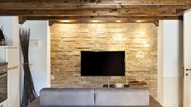 A modern living room with a stone wall, a flat-screen TV mounted above a sofa, and a wooden ceiling. - Baglivi 17 - Lecce Selection (Lecce)