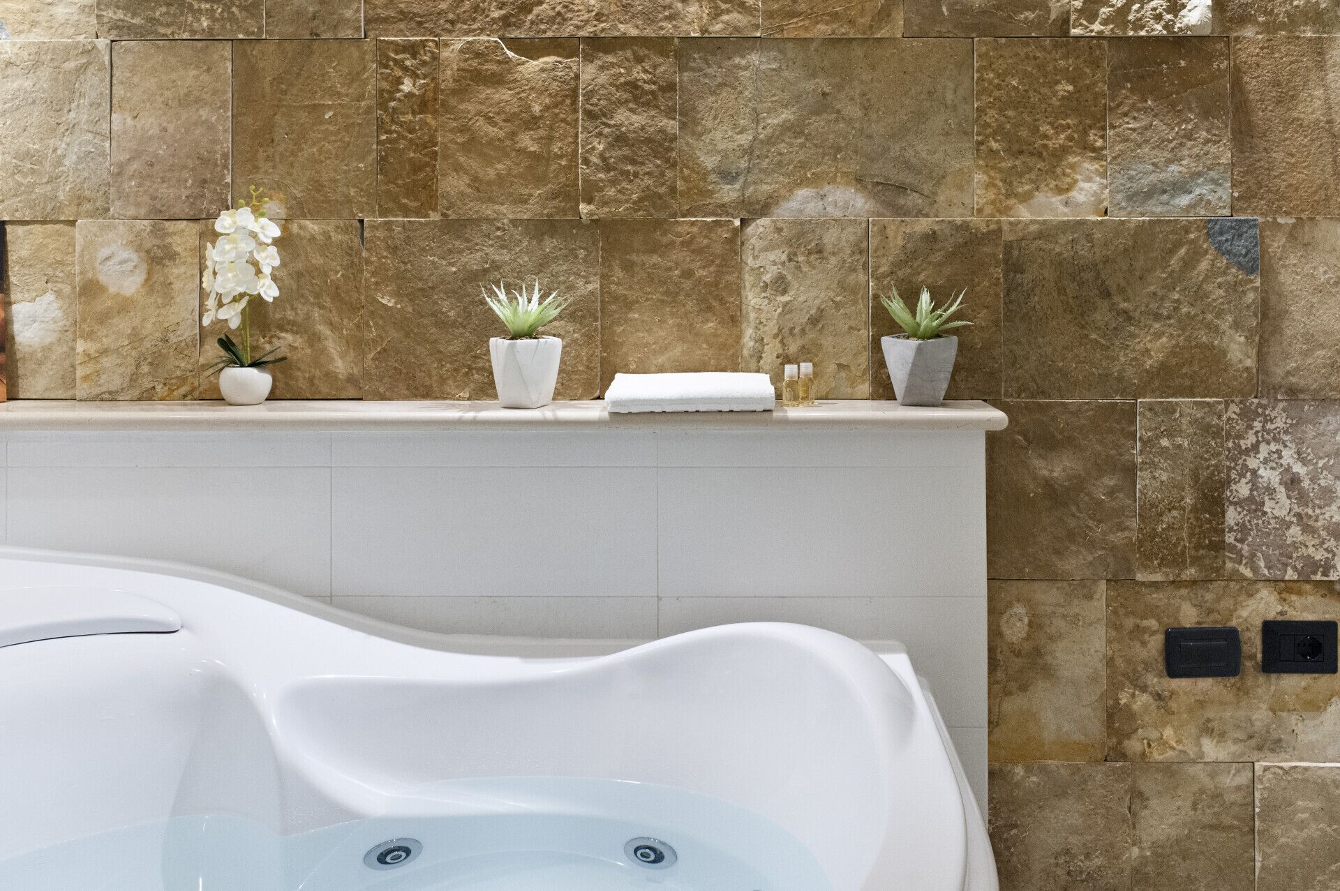A spa area with a white bathtub, stone wall tiles, and three potted plants on a ledge.
