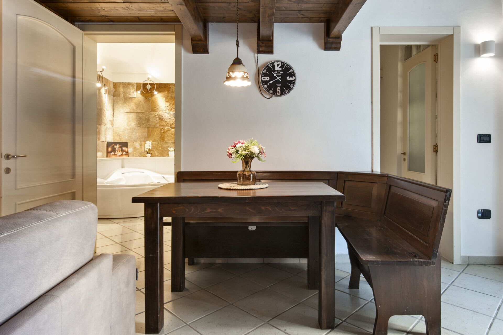 A dining area with a wooden table, bench seating, a wall clock, and a stone wall with a bathtub visible through an open door.