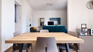 A modern living room with a wooden dining table, grey chairs, a blue sofa, and a TV mounted on the wall.