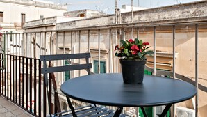 A balcony with a round table and a chair, a potted plant with red flowers, and a view of buildings and construction cranes in the background.