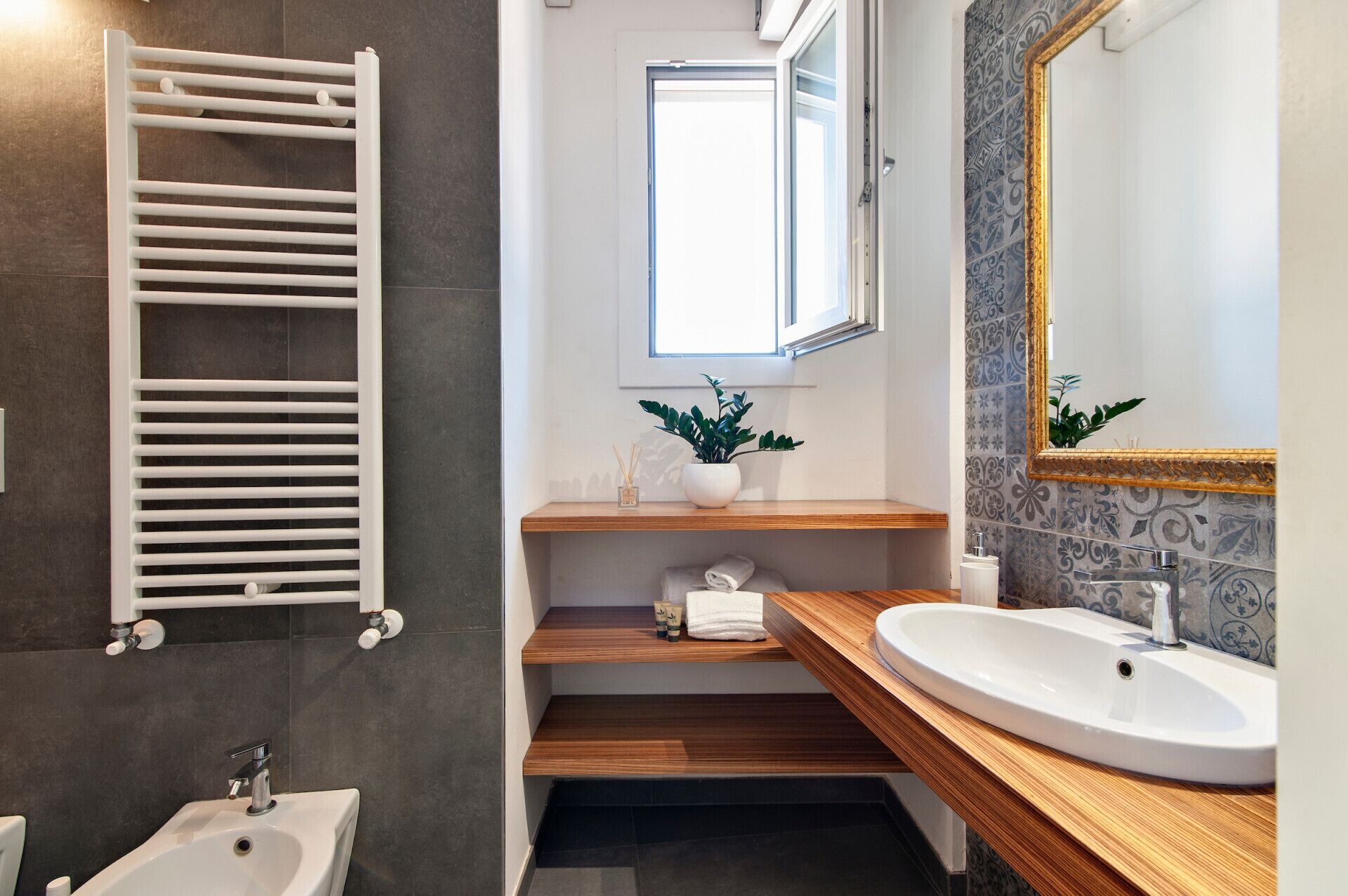 A modern bathroom with a white bathtub, a white sink, and a wooden vanity. There is a towel rack, a mirror with a decorative frame, and a window providing natural light.