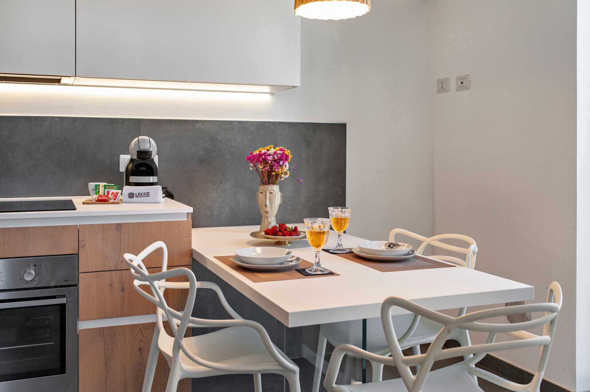 A modern kitchen with a coffee machine, a vase with flowers, and a dining table set for two with glasses of wine.