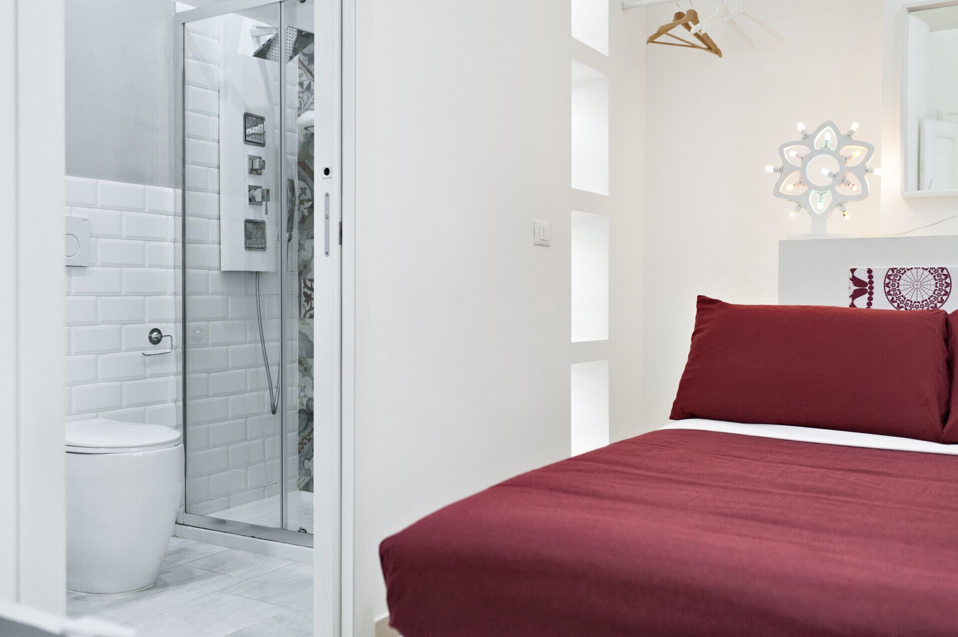 A modern bathroom with a glass shower enclosure, a white toilet, and a bed with a red bedspread.