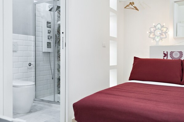 A modern bathroom with a glass shower enclosure, a white toilet, and a bed with a red bedspread.
