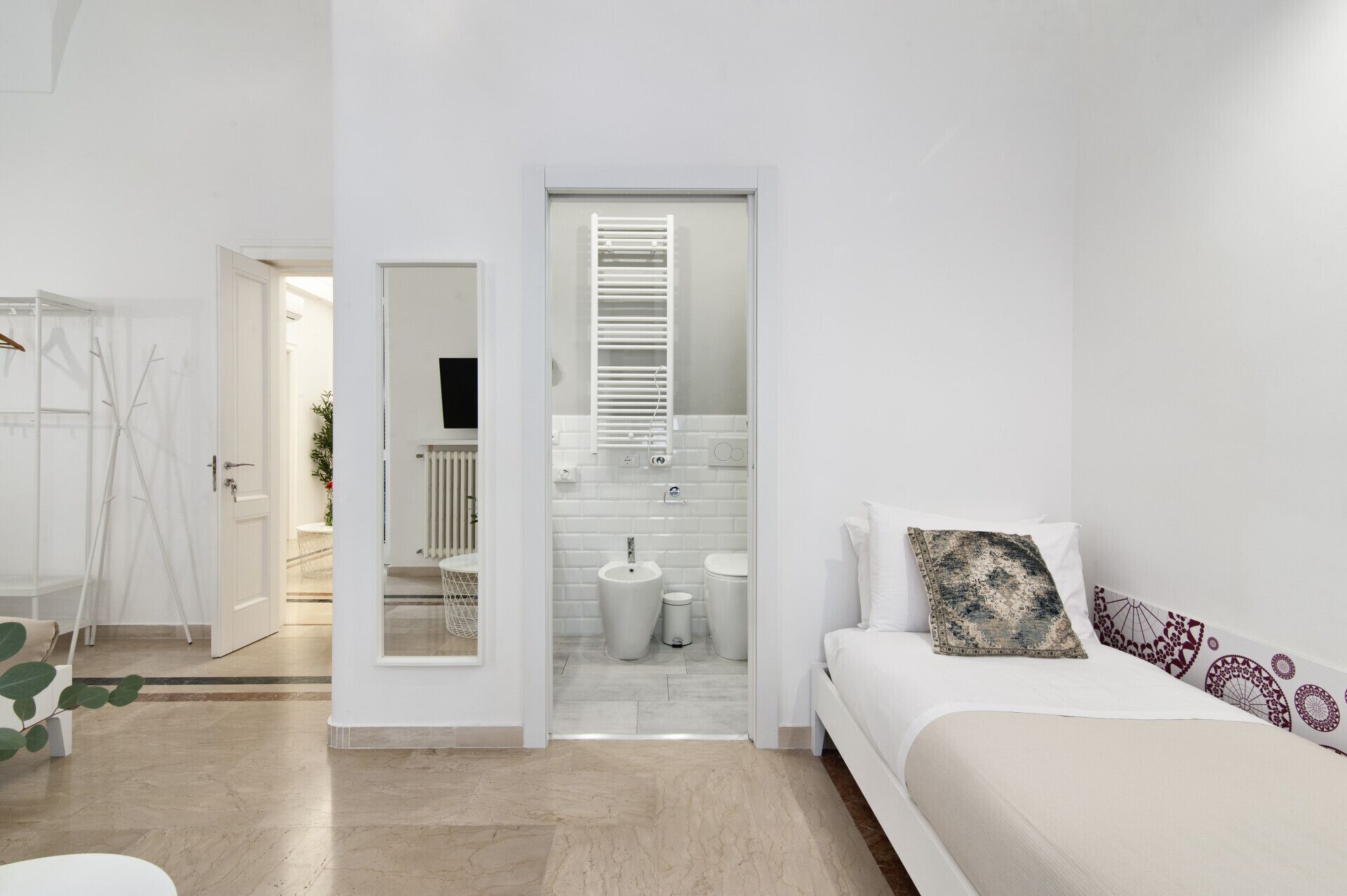 A modern bedroom with a bed, a bedside table, a mirror, and a bathroom visible through an open door.