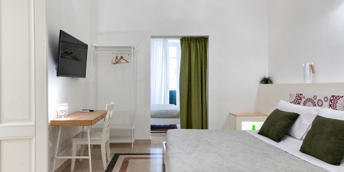 Une chambre à coucher comprenant un lit, un bureau, une chaise, un téléviseur fixé au mur et une fenêtre avec des rideaux.