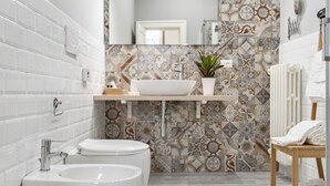 A modern bathroom with a white bathtub, a sink with a vessel basin, and a toilet. The walls are decorated with patterned tiles, and there is a small plant on the vanity.