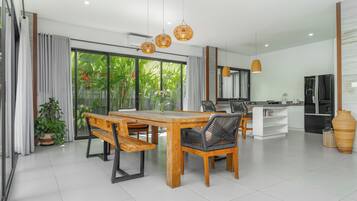 A modern dining area with a wooden table and benches, a kitchen in the background, and large windows with a view of greenery.