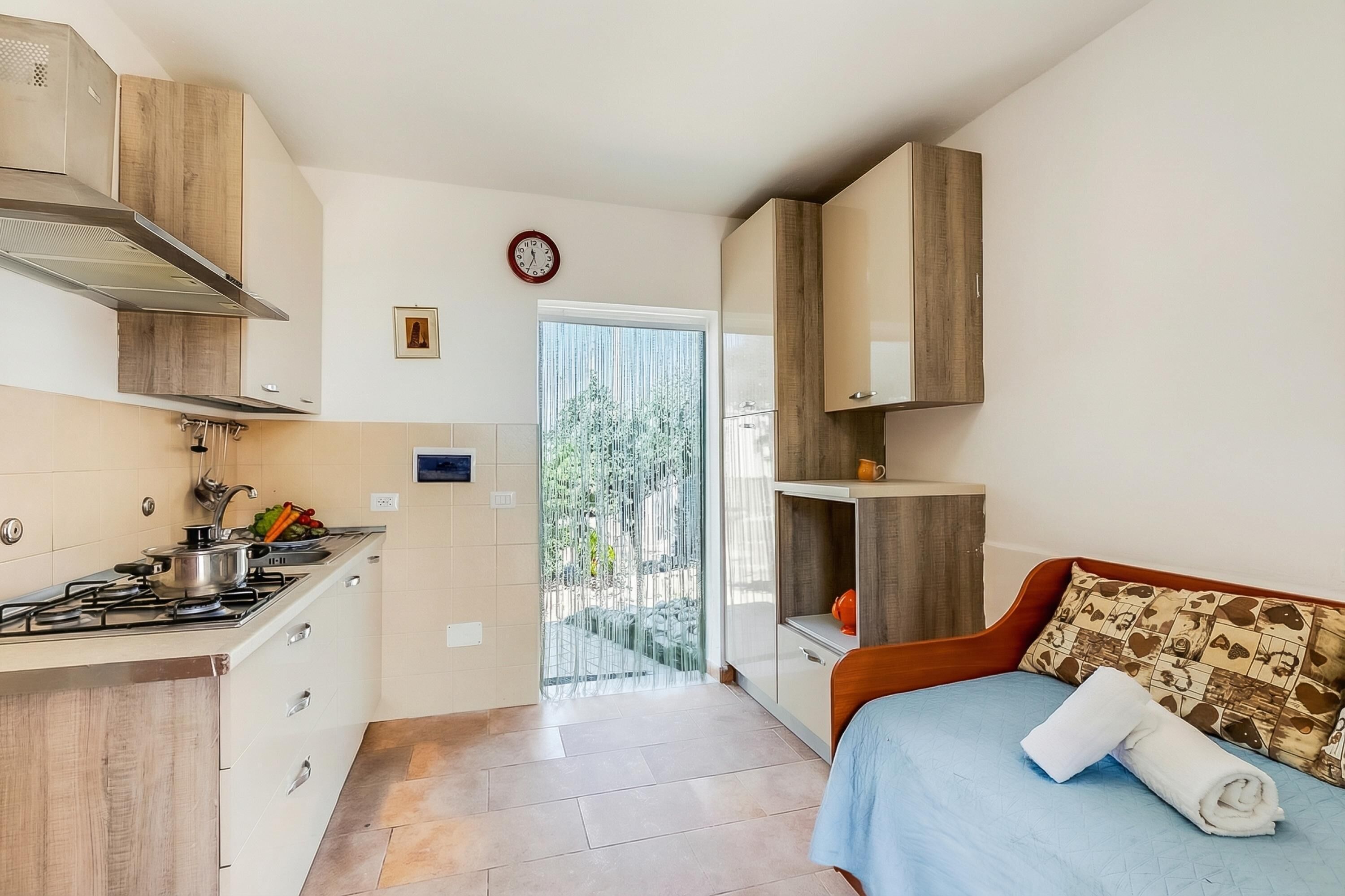 A compact hotel room with a kitchenette, a bed with a rolled towel, a clock, and a view through a glass door.
