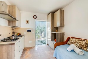 Una habitación de hotel compacta con kitchenette, una cama con una toalla enrollada, un reloj y vistas a través de una puerta de cristal.