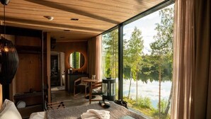 Un dormitorio con un ventanal grande con vista a un lago, techo de madera y una zona de estar acogedora con un espejo redondo.