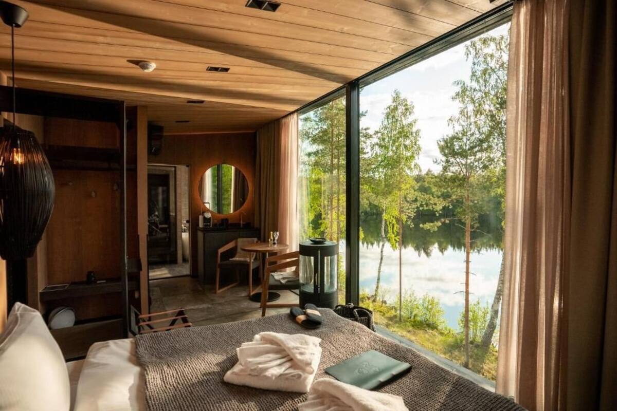 A bedroom with a large window overlooking a lake, a wooden ceiling, and a cozy seating area with a round mirror.