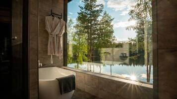 Un baño moderno con una bañera independiente, cabina de ducha con mampara de vidrio y vistas a un lago y árboles.