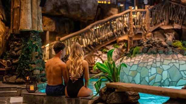 Una pareja sentada junto a una piscina, con un muro de piedra y un puente de madera al fondo.