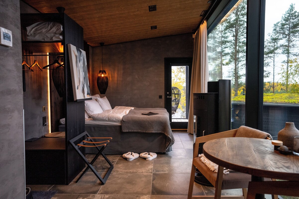 A modern bedroom with a bed, a wooden table, and a view of trees through a large window.