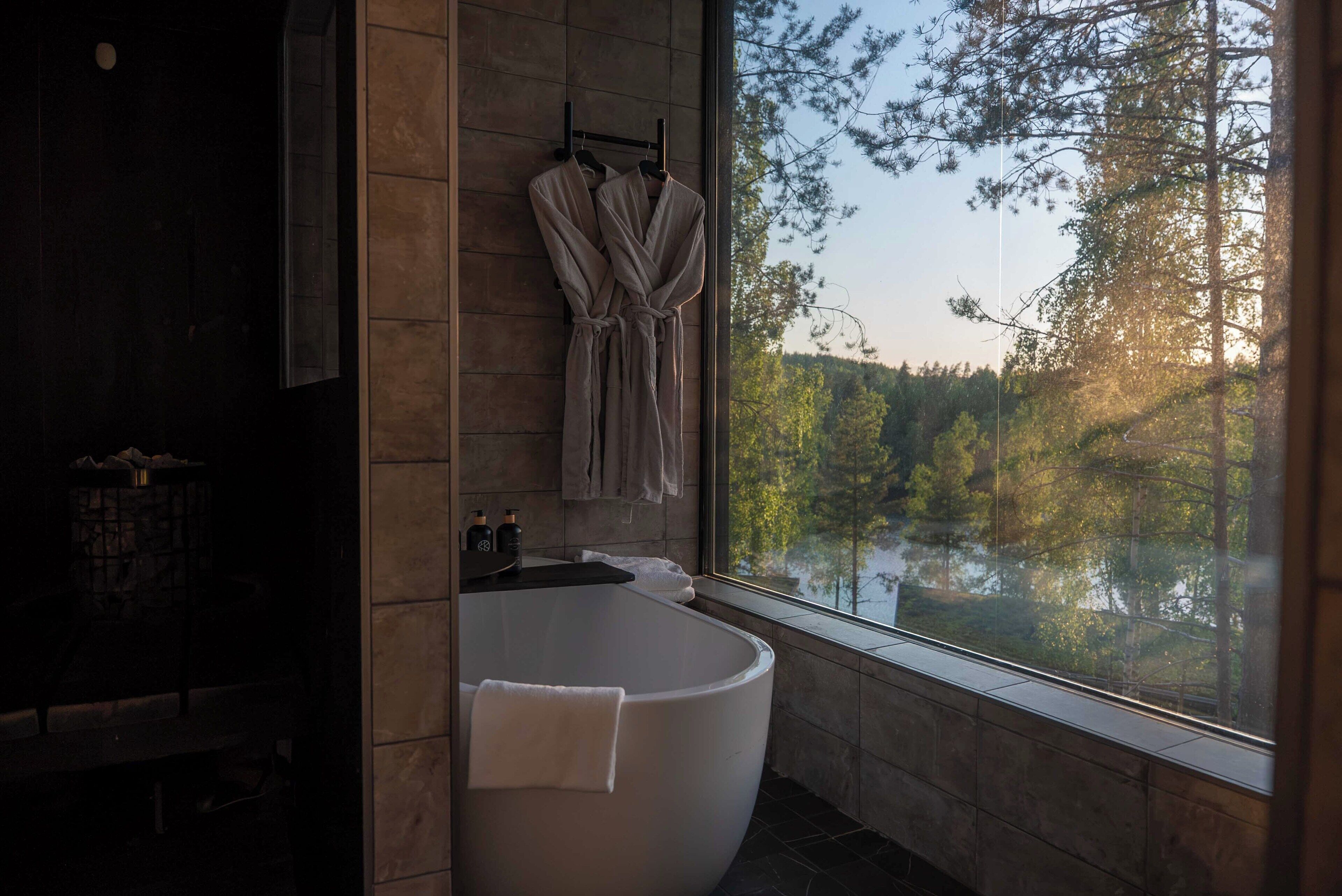 Une salle de bain moderne avec une baignoire sur pieds, une grande fenêtre donnant sur la verdure et un porte-serviettes avec des peignoirs.