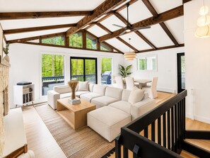 A spacious living area with a large sectional sofa, a wooden coffee table, and a dining table. There are multiple windows providing views of the outdoors.