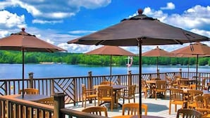 A lakeside patio with wooden tables and chairs, umbrellas, and a view of the lake. - Peaceful Two-Story Lake Naomi Home, Modern Design in Timber Trails + Lake Access (Pocono Pines)