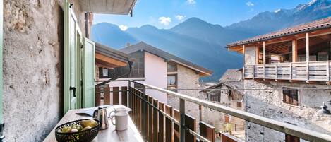 A balcony with a view of mountains, a wooden table with a basket of fruit, and a metal pitcher.