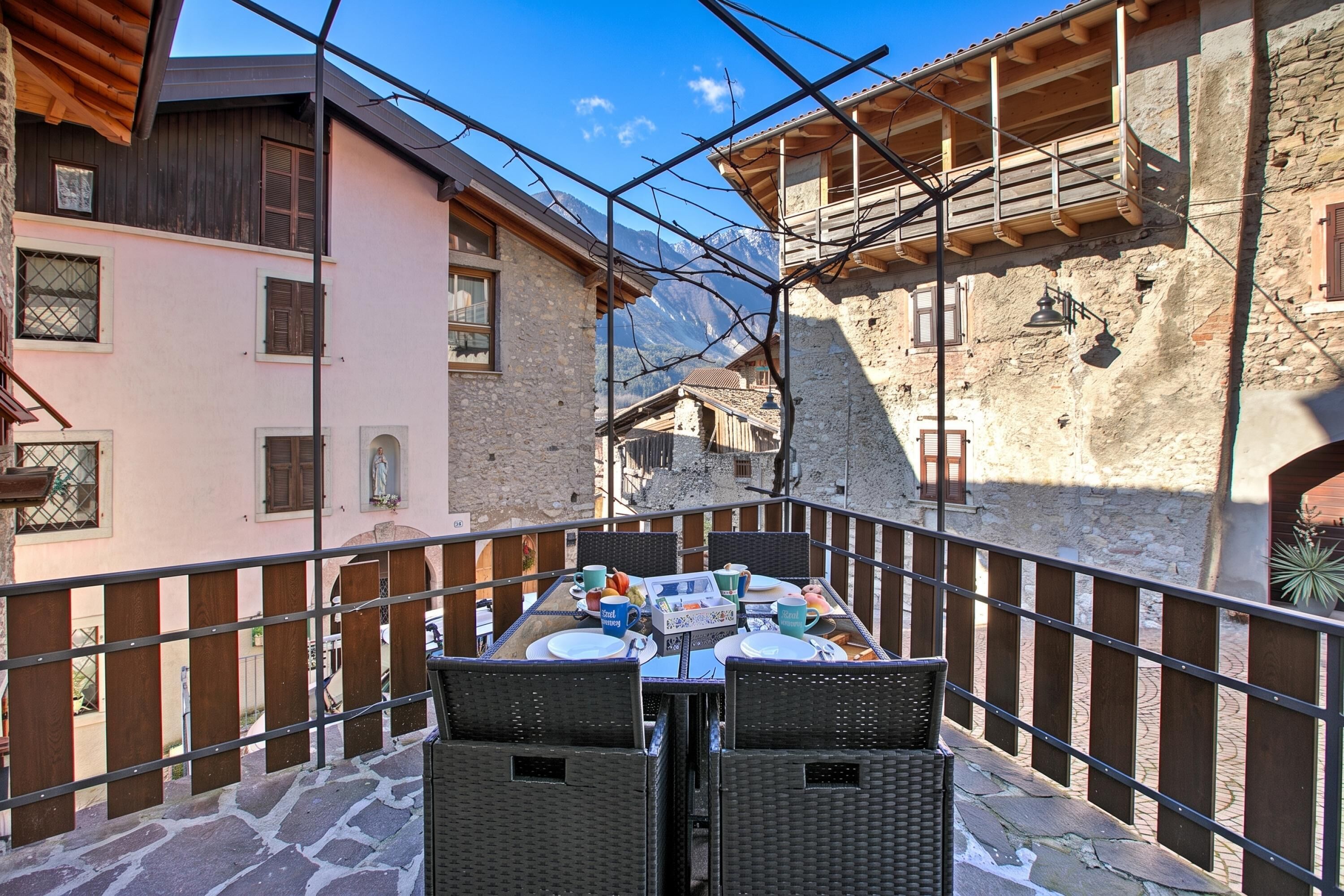 A balcony with wicker chairs and a table set for dining, overlooking a street with traditional buildings and a mountain in the background.