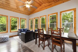 Une pièce spacieuse avec un plafond en bois, de grandes fenêtres, une table à manger avec des chaises, un canapé et un pouf à motifs.