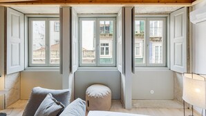 Une pièce avec un haut plafond en bois, un petit pouf, un canapé et trois fenêtres dont les volets sont ouverts.
