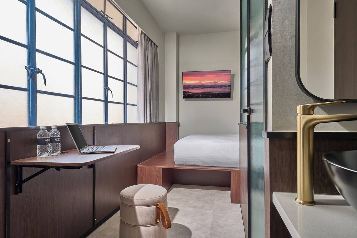 Une chambre d’hôtel compacte comprenant un lit, un bureau avec un ordinateur portable et des bouteilles d’eau, un petit tabouret et un tableau représentant un coucher de soleil accroché au mur.