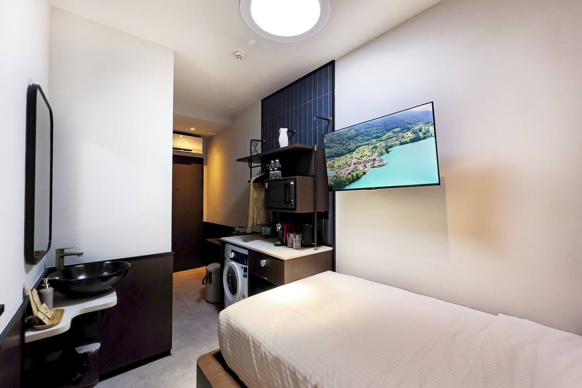 Une chambre d’hôtel moderne équipée d’un lit, d’une machine à laver, d’un lavabo, d’un miroir, d’une télévision et offrant une vue sur un paysage par la fenêtre.