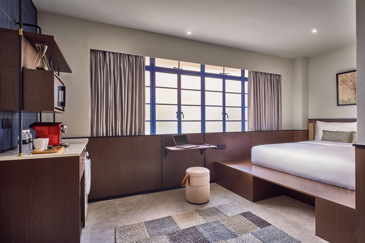 Une chambre d’hôtel moderne équipée d’un lit, d’un bureau avec un ordinateur portable, d’un petit tabouret, d’une machine à café et d’une grande fenêtre avec des rideaux.