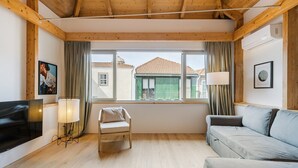 A cozy living room with a wooden ceiling, a large window with a view of buildings, a grey sofa, a white chair, and a flat-screen TV. - GuestReady - Cedofeita Collection Deluxe P3 (Porto)